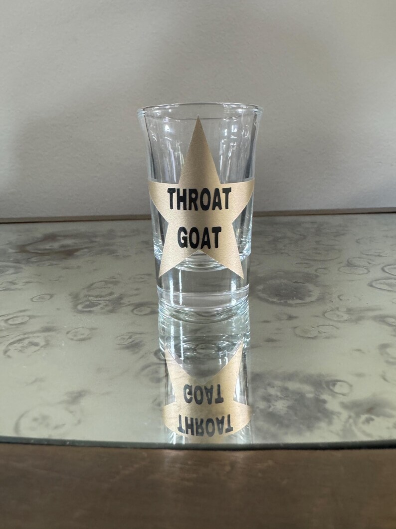May include: A clear glass shot glass with a gold star sticker that says "Throat Goat" on it. The shot glass is sitting on a table with a reflective surface.