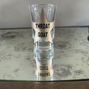 May include: A clear glass shot glass with a gold star sticker that says "Throat Goat" on it. The shot glass is sitting on a wooden surface.