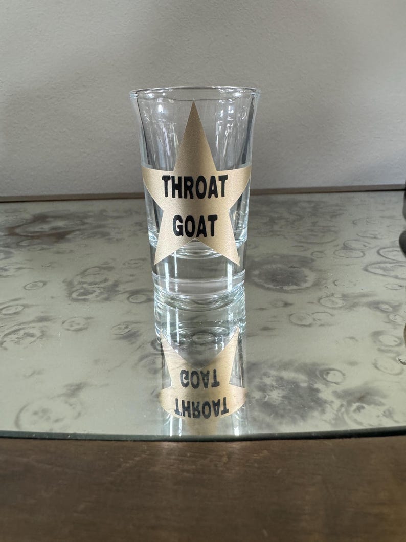 May include: A clear glass shot glass with a gold star sticker that says "Throat Goat" on it. The shot glass is sitting on a wooden surface.