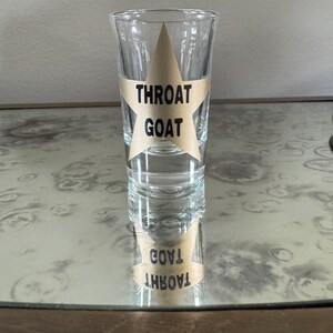 May include: A clear glass shot glass with a gold star sticker that says "Throat Goat" on it. The shot glass is sitting on a table.