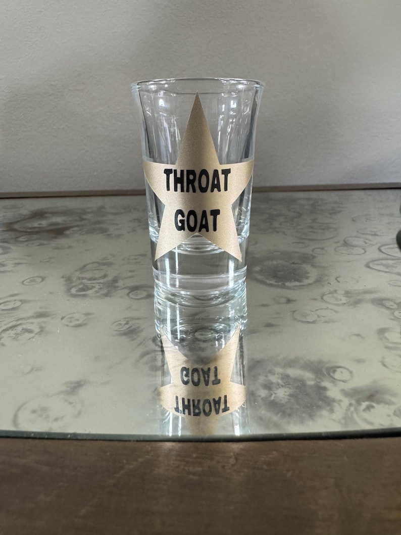 May include: A clear glass shot glass with a gold star sticker that says "Throat Goat" on it. The shot glass is sitting on a table.