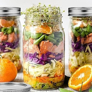 May include: Three glass mason jars filled with layered ingredients for a salad. The jars contain orange slices, green beans, red cabbage, and sprouts. The jars are on a white surface.