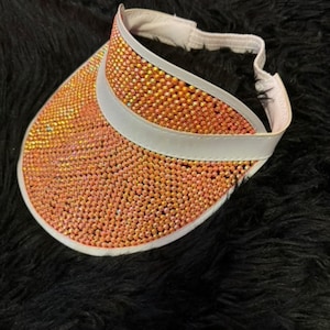 May include: A pink visor with a white band and a design of orange rhinestones.