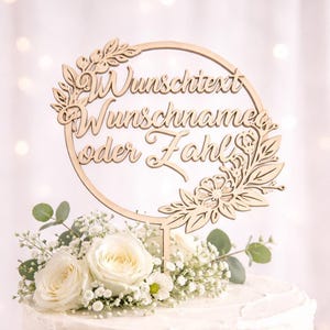 May include: A wooden cake topper with the words "Wunschtext Wunschname oder Fahl" in script, encircled by a floral wreath. The topper is placed on a white cake adorned with white roses and foliage.