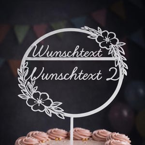 Könnte beinhalten: Ein weißer Tortenaufsatz mit den Worten "Wunschtext Wunschtext 2" in Schreibschrift. Der Aufsatz hat eine kreisförmige Form, verziert mit floralen und Blattmotiven. Der Aufsatz befindet sich auf einem Kuchen mit rosa Zuckerguss.