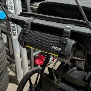 May include: A black stroller with a black and yellow Ryobi speaker attached. The stroller has a black canopy and a Thule storage bag. The wheels are black with red accents. The speaker is mounted on the stroller's handle.