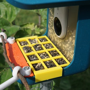 Pu&ograve; includere: Un mangiatoia per uccelli blu con un vassoio giallo contenente semi di girasole. La mangiatoia ha una telecamera incorporata e un posatoio per gli uccelli.