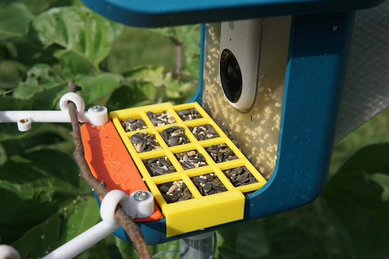 Pu&ograve; includere: Un mangiatoia per uccelli blu con un vassoio giallo contenente semi di girasole. La mangiatoia ha una telecamera incorporata e un posatoio per gli uccelli.