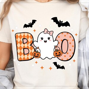 Può includere: T-shirt color crema con grafica a tema Halloween. Il design presenta la parola "BOO" con un personaggio fantasma che tiene in mano delle zucche, circondato da pipistrelli e stelle. La lettera "B" ha un motivo a rombi arancione e bianco, e la "O" ha un motivo a pois rosa.