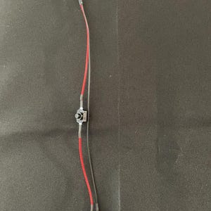 May include: A small electronic component with a yellow LED light at the top. The component has red and clear wires, a small black switch, and a black battery connector at the bottom. The background is a dark, textured surface.