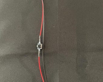 May include: A small electronic component with a yellow LED light at the top. The component has red and clear wires, a small black switch, and a black battery connector at the bottom. The background is a dark, textured surface.