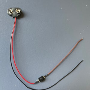 Puede incluir: Un paquete de baterías negro con cables rojo y negro conectados a un interruptor de botón pulsador negro.