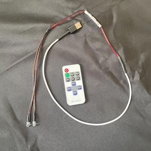 Könnte beinhalten: Ein weißer, USB-betriebener LED-Lichtstreifen mit zwei klaren LED-Birnen an den Enden dünner schwarzer und roter Kabel. Das Gerät enthält eine kleine weiße Fernbedienung mit einem roten Power-Knopf, grauen Funktionstasten und dem Text RF Wireless.