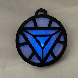 Puede incluir: Un colgante impreso en 3D en blanco y negro con el diseño del reactor de arco de Iron Man. El reactor de arco brilla en azul.