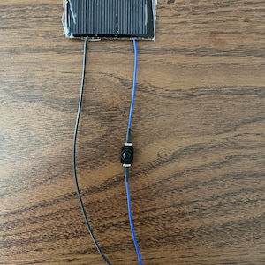 Könnte beinhalten: Ein kleines Solarpanel mit zwei daran befestigten Drähten. Ein Draht ist schwarz und der andere blau. Die Drähte sind mit einem kleinen schwarzen Schalter verbunden.