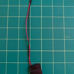 May include: A small electronic component featuring a black plastic battery holder connected by one red and one black wire to a single blue LED light bulb. The wires are thin and flexible, extending from the battery pack to the light source.
