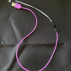 Può includere: Un cavo USB viola con un connettore USB-A e una serie di fili con piccole luci LED all'estremità. Il cavo è su una superficie nera.