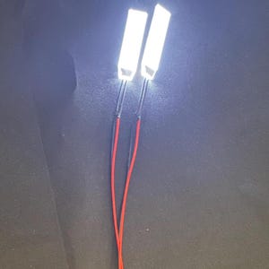 Può includere: Due strisce luminose a LED bianche e luminose con fili rossi e neri. Le luci rettangolari sono illuminate, emettendo una forte luce focalizzata. I fili si estendono verso il basso, suggerendo una connessione o una fonte di alimentazione.