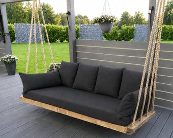 Può includere: Un dondolo da veranda rettangolare con una struttura in legno naturale sospesa da spesse corde intrecciate di colore chiaro. Il dondolo include un lungo cuscino di seduta grigio scuro e quattro cuscini quadrati abbinati per lo schienale.