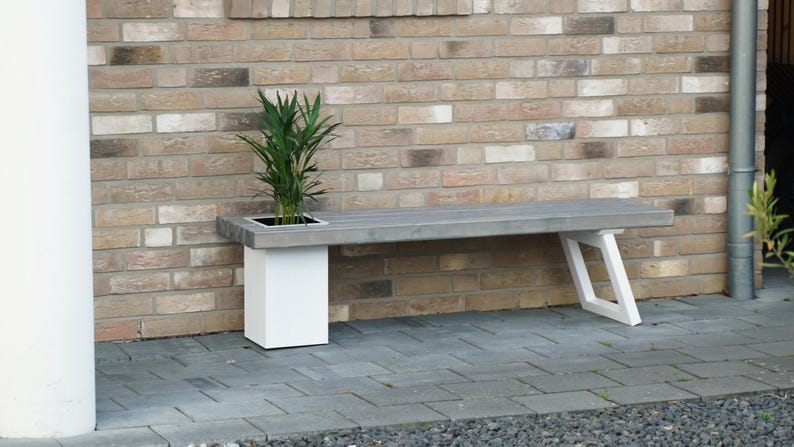 May include: A white wooden bench with a built-in planter, featuring a green plant. The bench is set against a brick wall.