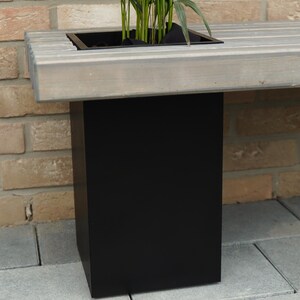 May include: A wooden bench with a built-in planter box. The planter box is made of black metal and is filled with green plants. The bench is sitting on a brick patio.