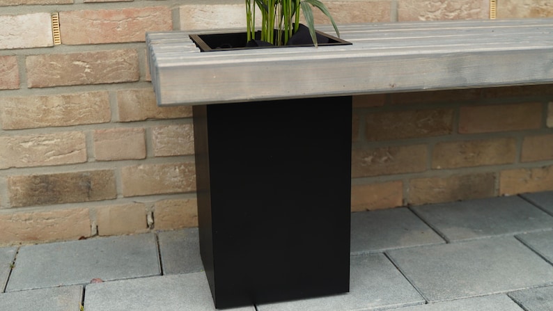 May include: A wooden bench with a built-in planter box. The planter box is made of black metal and is filled with green plants. The bench is sitting on a brick patio.