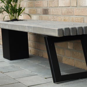 May include: A concrete bench with a gray top and black metal legs. The bench has a unique design with a zig-zag pattern on the underside of the top.