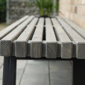 May include: A close-up of a gray wooden bench with black metal legs. The bench is made of slats of wood and has a simple, modern design.
