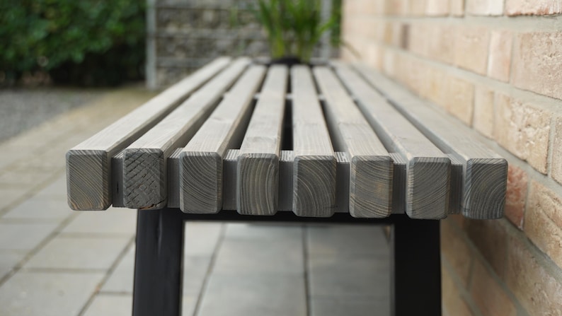 May include: A close-up of a gray wooden bench with black metal legs. The bench is made of slats of wood and has a simple, modern design.