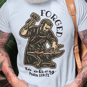May include: Light blue t-shirt featuring a blacksmith graphic with the words "FORGED to obey Psalm 119:71". The illustration depicts a blacksmith working at an anvil. The shirt is made of a soft, comfortable material.