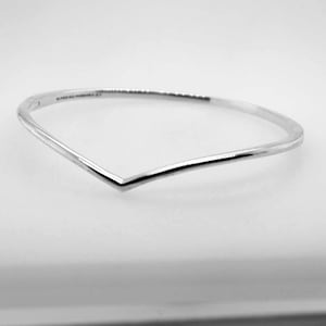 May include: A silver-coloured, V-shaped bangle bracelet. The bracelet has a minimalist design with a smooth, polished finish. The bracelet is a piece of jewellery.