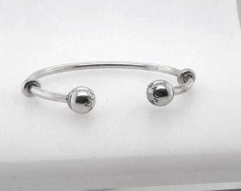 May include: A silver-colored open bangle bracelet. The bracelet has a curved design with a ball at each end. Each ball has a small design etched into it. The bracelet is displayed on a white surface.