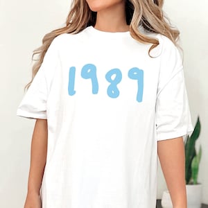 May include: White oversized t-shirt with a light blue graphic of the year 1989.