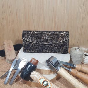 Op de afbeelding: Een zwarte leren portemonnee met een slangenhuidpatroon en gouden stiksels. De portemonnee is omringd door leerbewerkingstools, waaronder een hamer, een priem en een roltool.