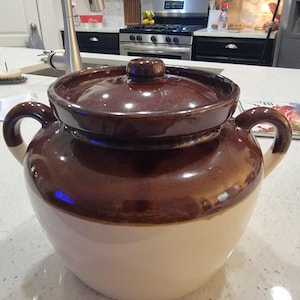 May include: A two-tone ceramic crock pot with a lid. The upper portion and lid are a dark brown, while the lower portion is cream-colored. It has two side handles and is likely used for cooking.