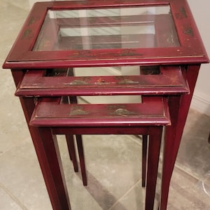 May include: Set of three nesting tables in a deep red hue. Each table features a glass top and decorative painted accents. The tables have slender legs and a classic design, suitable for various interior styles.