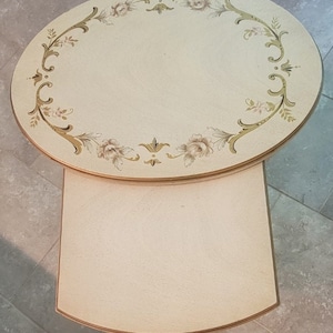 May include: Cream-colored oval side table with a lower rectangular shelf. The tabletop features a floral design in gold and muted pink. The table has a classic, vintage aesthetic.