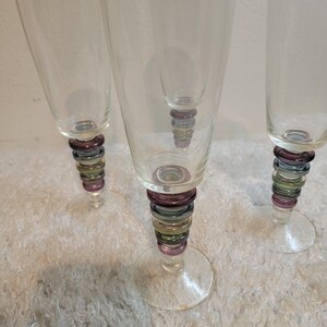 May include: Set of three clear champagne flutes. Each glass features a stem adorned with stacked, colorful rings in shades of purple, green, and gold. The glasses have a classic, elegant design.
