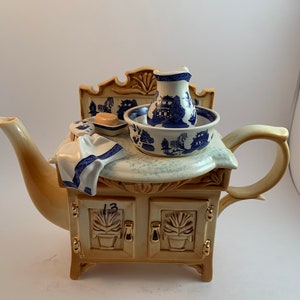 May include: A ceramic teapot shaped like a vanity with a blue and white pitcher, bowl, and soap dish. The teapot is decorated with gold accents and has a number "13" on the front.