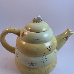 Op de afbeelding: Een gele theepot van keramiek in de vorm van een bijenkorf met een bij op de deksel. De theepot heeft een handvat en een tuit. De theepot is versierd met bijen en blauwe stippen.