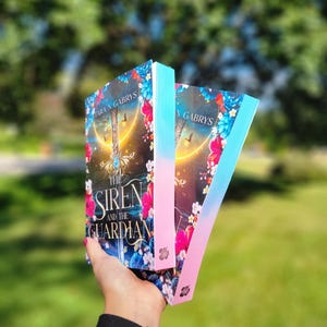 Könnte beinhalten: Zwei Exemplare des Buches "The Siren and the Guardian" werden vor einem verschwommenen grünen Hintergrund gehalten. Die Buchcover zeigen eine Fantasy-Szene mit einem Schwert, Blumen und dem Namen des Autors. Die Buchkanten sind in einem Rosa-Blau-Verlauf bemalt.