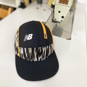 Op de afbeelding: Zwarte New Balance pet met een gebogen klep. De pet heeft een wit logo en een band met een patroon van bruine, zwarte en witte strepen. Gele biezen accentueren het ontwerp. Waarschijnlijk gemaakt van lichtgewicht, ademend materiaal.