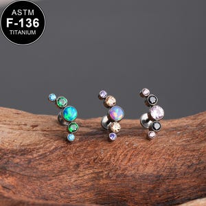 May include: Three titanium ear piercings with curved designs. Each features a cluster of colorful, iridescent gemstones in shades of blue, green, purple, pink, and black. The piercings are displayed on a wooden surface. The text "ASTM F-136 TITANIUM" is in the upper left corner.