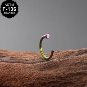 Peut inclure: Un anneau de nez incurvé en titane. L'anneau présente une finition arc-en-ciel avec un accent rose. L'image comprend un cercle noir avec le texte "ASTM F-136 TITANIUM". L'anneau est posé sur un fond en bois.