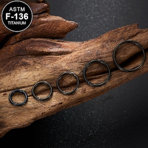 May include: Five black titanium hinged segment rings of varying sizes are displayed on a piece of dark, textured wood. The rings are arranged from smallest to largest. The top left corner of the image features a circular logo with the text "ASTM F-136 TITANIUM".