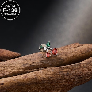May include: A silver-colored titanium earring with a cherry design. The cherry features a red heart-shaped gem, a small red gem, and a green leaf-shaped gem. The earring has a flat back and a small, round, white gem. The text "ASTM F-136 TITANIUM" is in a white circle.