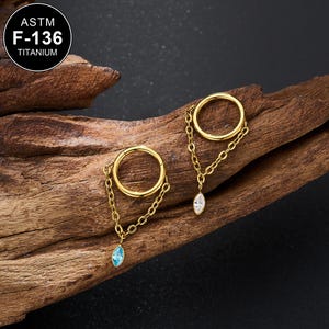 May include: Two gold-colored hoop earrings with dangling chains and gemstones. One earring features a blue gemstone, the other a clear gemstone. The earrings are made of titanium and are displayed on a piece of wood. The text "ASTM F-136 TITANIUM" is visible.