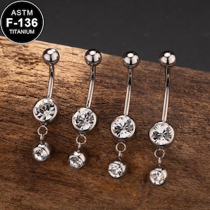 Puede incluir: Cuatro anillos de ombligo plateados con colgantes de cristal transparente. Los anillos están hechos de titanio ASTM F-136.