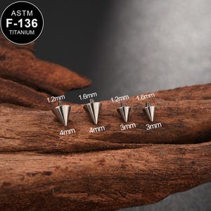 Puede incluir: Cuatro pendientes de joyería corporal plateados en forma de cono con respaldos de rosca. Los pendientes están etiquetados con medidas: 1,2 mm, 1,6 mm, 4 mm y 3 mm. La esquina superior izquierda tiene un logotipo circular con el texto "ASTM F-136 TITANIUM".