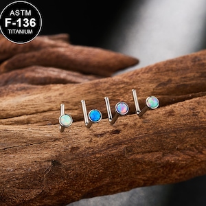 Könnte beinhalten: Vier kleine, silberfarbene Nasenstecker mit opalartigen, runden Edelsteinen in verschiedenen Farben. Die Stecker sind auf einem Holzstück in einer Reihe angeordnet. In der oberen linken Ecke des Bildes befindet sich ein rundes Logo mit dem Text "ASTM F-136 TITANIUM".